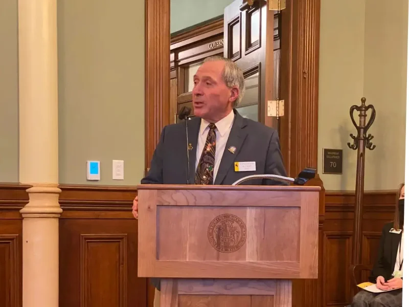 RJ Kost speaking at a podium in a government building, demonstrating leadership and public service